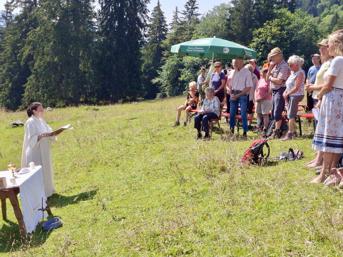 Auf der grünen Schräge einer Almwiese stehen rechts erhöht andachtsvolle Gottesdienstbesucher*innen, die sich von roten Bierbänken erhoben haben. Auch ein grüner Sonnenschirm ist zu sehen. Links, auf der Talseite, ist mit einem Holztisch ein Feldaltar hergerichtet. Davor steht eine Frau in weißem, langem Gewand, die eine Mappe aufgeschlagen hat und zu den  Gottesdienstbesucher*innen spricht. Im Hintergrund wird die Wiese von dunkelgrünem Wald begrenzt. Ein bisschen blauer Himmel ist auch zu sehen.