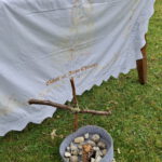 Nahaufnahme des Altars aus dem vorigen Bild: Über den Tisch ist ein weißes, besticktes Altartuch mit gezackten Rändern gelegt, das fast bis auf den Wiesenboden reicht. Gestickt ist in Gelb/Gold und Rot. Zu erkennen ist ein Christus-Kopfporträt und darunter der Text „Gelobt sei Jesus Christus“. Davor, auf dem Boden, ist ein kleines, aus abgebrochenen Zweigen geschnürtes Holzkreuz gesteckt, vor dem wiederum in einem grauen Filzkorb gesammelte Steine liegen.