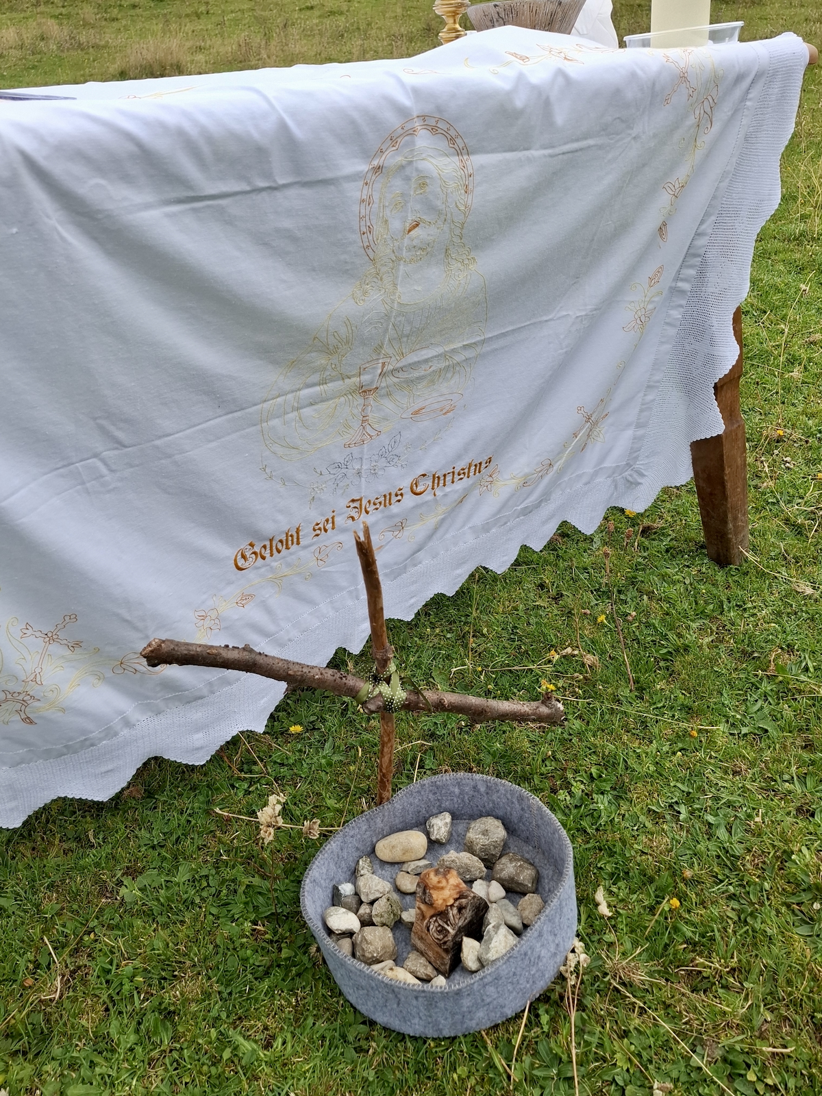 Nahaufnahme des Altars aus dem vorigen Bild: Über den Tisch ist ein weißes, besticktes Altartuch mit gezackten Rändern gelegt, das fast bis auf den Wiesenboden reicht. Gestickt ist in Gelb/Gold und Rot. Zu erkennen ist ein Christus-Kopfporträt und darunter der Text „Gelobt sei Jesus Christus“. Davor, auf dem Boden, ist ein kleines, aus abgebrochenen Zweigen geschnürtes Holzkreuz gesteckt, vor dem wiederum in einem grauen Filzkorb gesammelte Steine liegen. 
