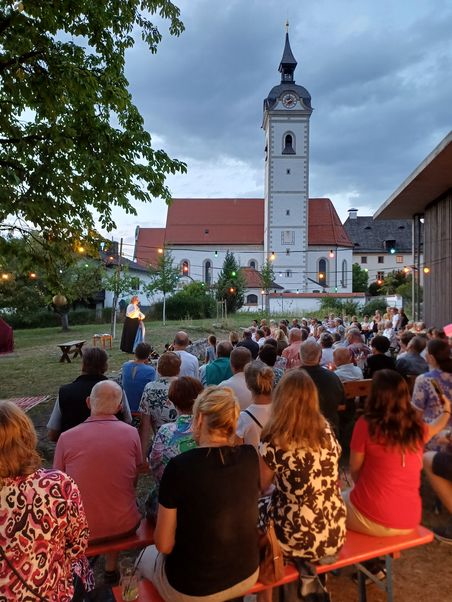 Foto in der Dämmerung: Eine Menge Menschen sitzt auf Bierbänken und hat den Blick auf eine etwas erhöhte Grasfläche mit jungen Obstbäumen gerichtet. Dort ist eine Frau mit dunklem Rock und weißer Bluse beleuchtet. Über dem Publikum sind Schnüre mit bunten Glühbirnen gezogen. Von links ragt ein belaubter Ast ins Bild. Im Hintergrund ist eine Kirche mit Turm zu sehen. Am Himmel sind deutliche graue Wolken.