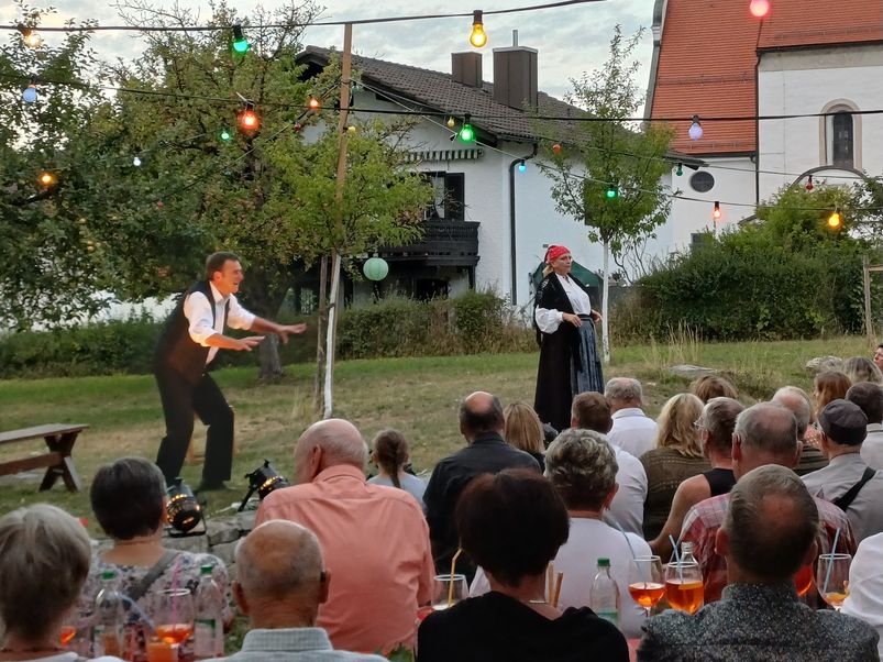 Foto: Blick über die Köpfe des Publikums hinweg auf eine Wiese mit jungen Obstbäumen; einige orange Cocktails sind zu sehen. Auf der Wiese spielen zwei Personen Theater: eine Frau mit dunklem Rock, weißer Bluse, schwarzem Umhang und roter Haube sowie ein Mann mit weißem Hemd und dunkler Hose sowie Weste. Im Hintergrund sind Häuser zu sehen, über den Menschen hängen bunt leuchtende Glühbirnen.