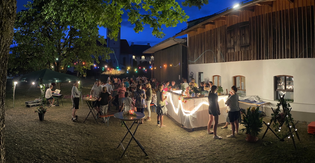 Nachtaufnahme: Im Freien vor einem historischen Stallgebäude, das unten gemauert und oben aus Holz ist, ist eine provisorische Bartheke aufgebaut, die mit Lichtschlangen erleuchtet ist. Drumherum und an Stehtischen stehen Leute und unterhalten sich, über ihnen leuchten sind bunte Glühbirnen. Unter einem Pavillon links spielt ein Mann auf einem Keyboard. Von links ragt ein Laubbaum ins Bild, ein weiterer Baum steht weiter hinten. Ganz hinten ist vor nachtblauem Himmel die Silhouette einer Kirche zu erahnen.