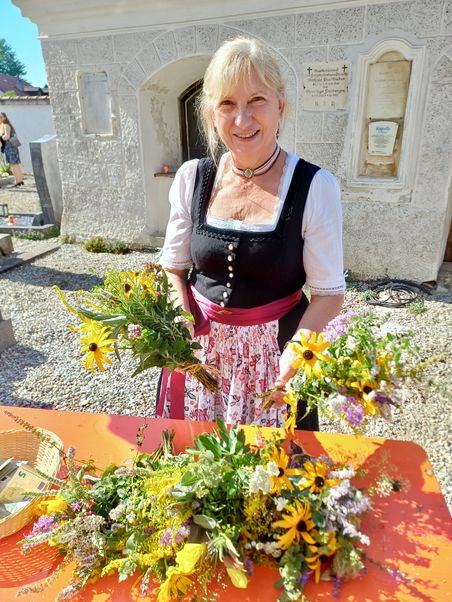 Foto, außen: Hinter einem roten Biertisch steht eine blonde Frau in Dirndltracht, die freundlich in die Kamera blickt und zwei Kräuterbuschn in den Händen hält. Auf dem Tisch liegen weitere Kräutersträuße. Angeschnitten ist auch ein Körbchen mit Geld zu sehen. Im Hintergrund ist ein helles, niedriges Kapellengebäude zu sehen, ein Grabstein lässt erahnen, dass der Ort ein Kirchfriedhof ist.