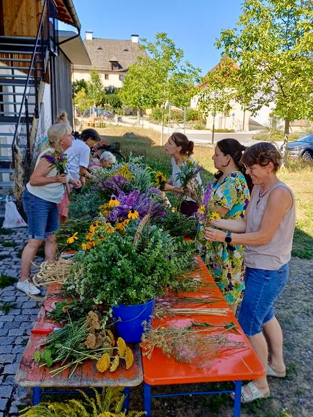 Vor einem Gebäude mit metallener Außentreppe sind zwei Biertische aneinandergerückt, sodass sie eine Arbeitsfläche bilden, an der eine mehrere Frauen von rechts und links Kräuterbuschn binden. Die Blumen und Kräuter stehen in Töpfen und Eimern auf den Tischen. Es ist ein sonniger Tag, im Hintergrund sind Häuser und eine Grasfläche mit jungen Bäumen zu sehen.