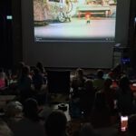 Foto: Blick aus einem dunklen Saal auf eine Kinoleinwand. Dort läuft ein Film mit der Zeichentrickfigur Pumuckl. Die Zuschauer*innen sind offenbar Kinder, sie haben sich auf den B0oden gekauert, ein einzelner unbesetzter Stuhl ist zu erkennen.