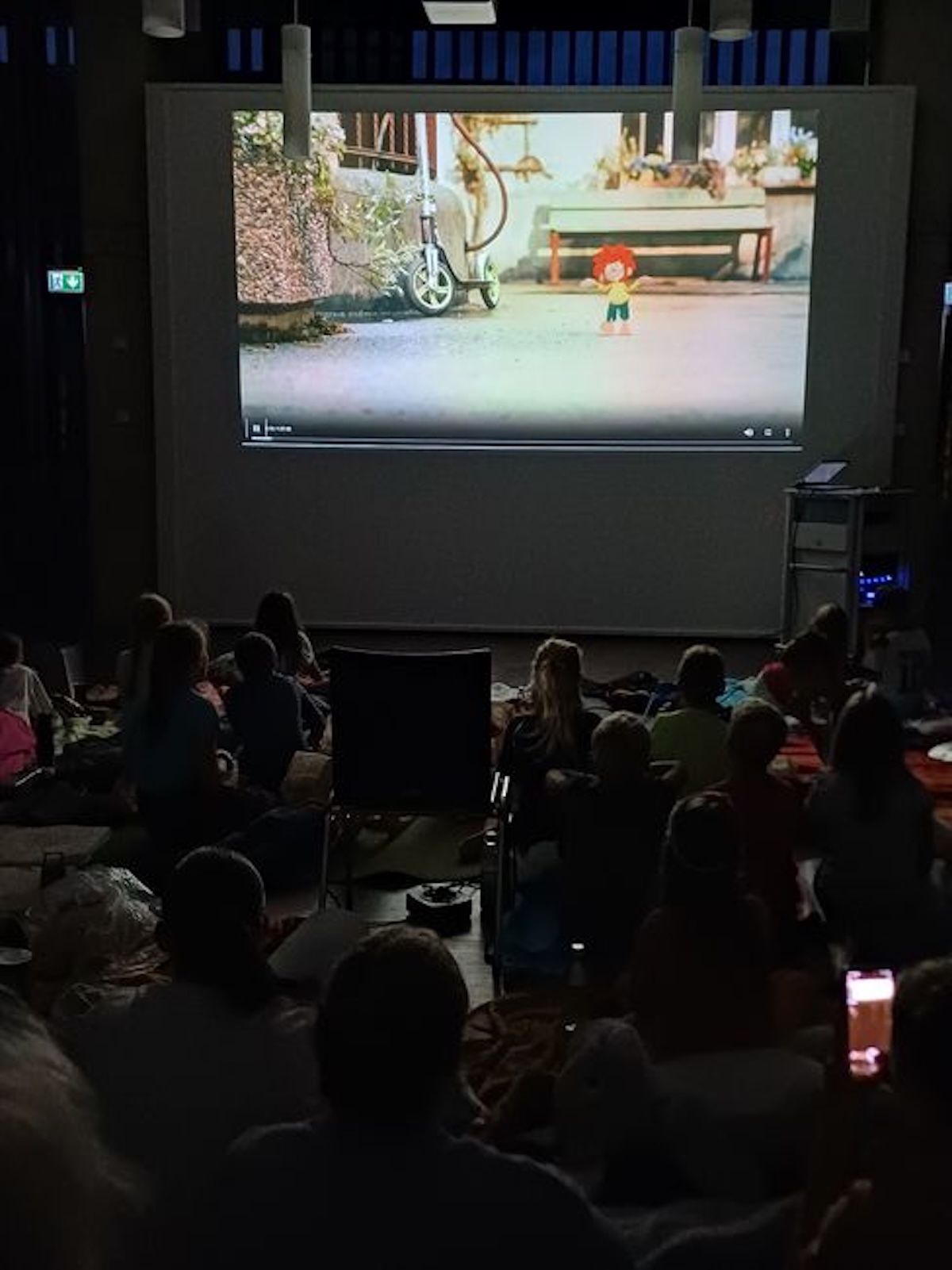 Foto: Blick aus einem dunklen Saal auf eine Kinoleinwand. Dort läuft ein Film mit der Zeichentrickfigur Pumuckl. Die Zuschauer*innen sind offenbar Kinder, sie haben sich auf den B0oden gekauert, ein einzelner unbesetzter Stuhl ist zu erkennen.