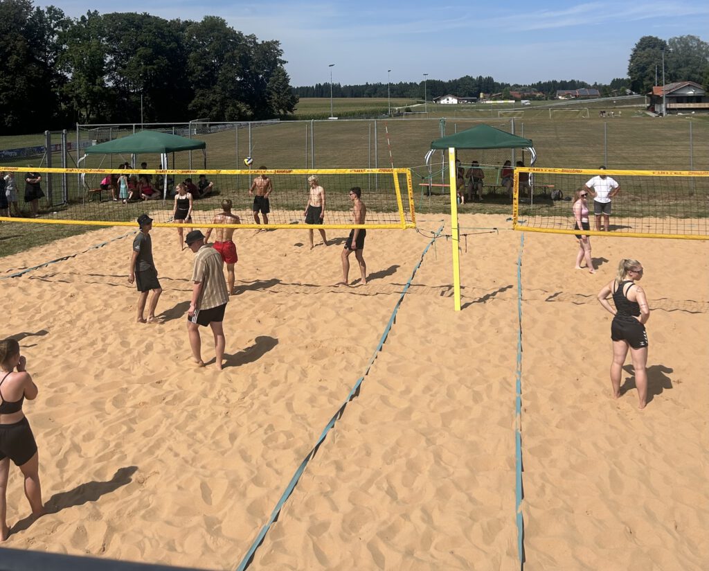 Blick von erhöhter Position auf ein Beachvolleyballgelände mit mit zwei Netzen. Das Gelände ist hoch eingezäunt, auf dem Gras um die Felder sind grüne Pavillons aufgestellt, unter denen Leute Schatten finden. Auf dem hellen Sand der Spielfelder stehen abwartend Leute in sommerlicher Bade- und Freizeitkleidung. Im Hintergrund sind ein Fußballfeld und einzelne Gebäude vor einem Waldsaum zu sehen. 