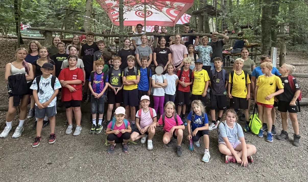 Gruppenbild von jungen Leuten und Kindern in sommerlich-sportlicher Freizeitkleidung auf einem gekiesten Bereich in einem schattigen Waldgelände. Im Hintergrund Bäume und eine Art Podium mit Bierbänken und Langnese-Sonnenschirmen.