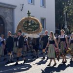 Auf der Straße vor einem Stadthaus zieht die Landjugend vorüber. Vier junge Männer in Tracht tragen auf Stangen eine Erntekrone. Daneben gehen Reihen von jungen Frauen in Dirndltracht mit hellgrünen Schürzen und rosa Bändern, die eine Blumengirlande tragen. Auf dem Gehweg dahinter begleiten Passanten den Zug. Im Hintergrund sind Laubbäume. Das Gegenlicht gibt der Ährenkrone starken Kontrast, der Zug wirft deutliche Schatten auf dem Asphalt voraus.