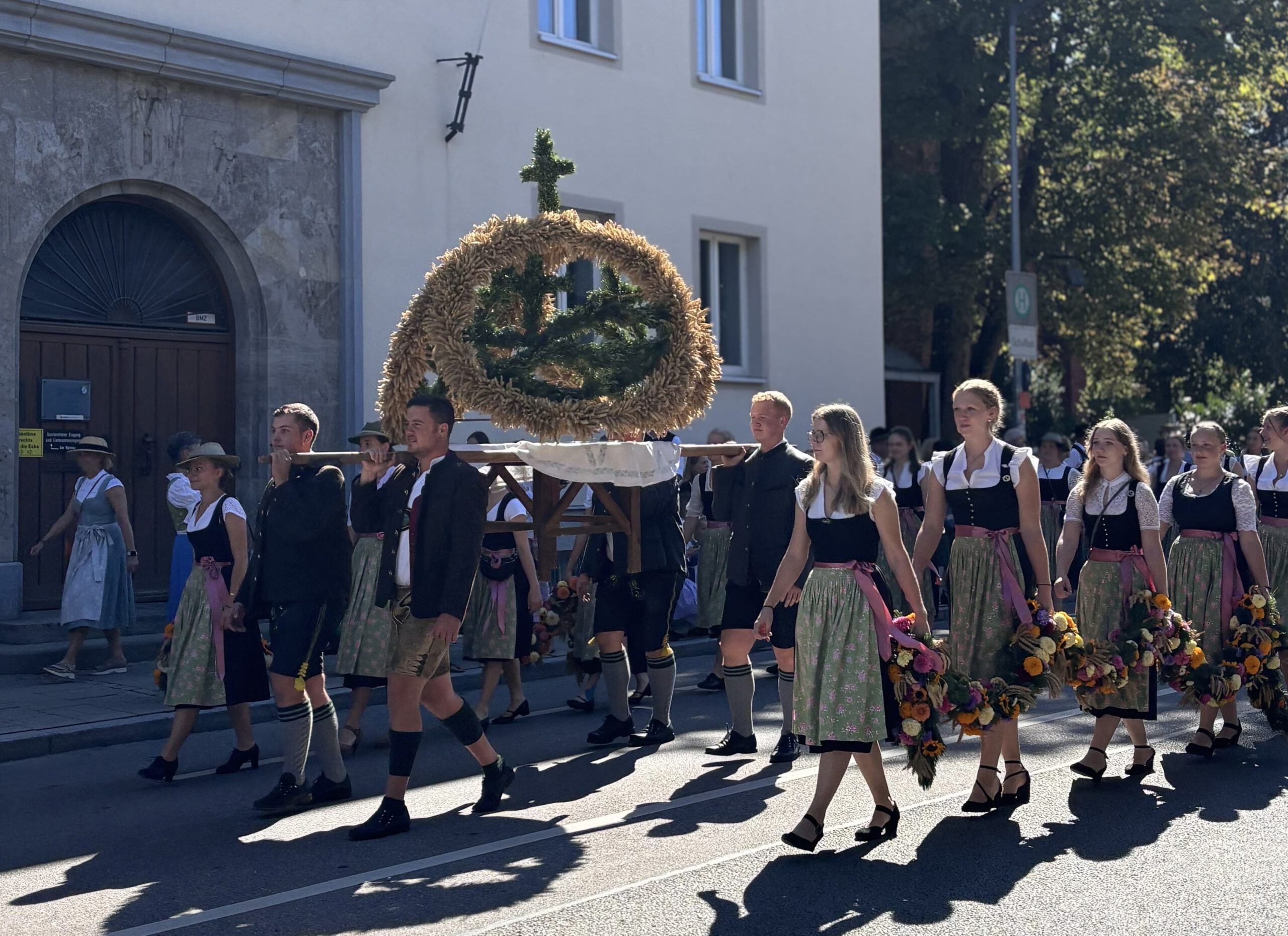 Auf der Straße vor einem Stadthaus zieht die Landjugend vorüber. Vier junge Männer in Tracht tragen auf Stangen eine Erntekrone. Daneben gehen Reihen von jungen Frauen in Dirndltracht mit hellgrünen Schürzen und rosa Bändern, die eine Blumengirlande tragen. Auf dem Gehweg dahinter begleiten Passanten den Zug. Im Hintergrund sind Laubbäume. Das Gegenlicht gibt der Ährenkrone starken Kontrast, der Zug wirft deutliche Schatten auf dem Asphalt voraus.