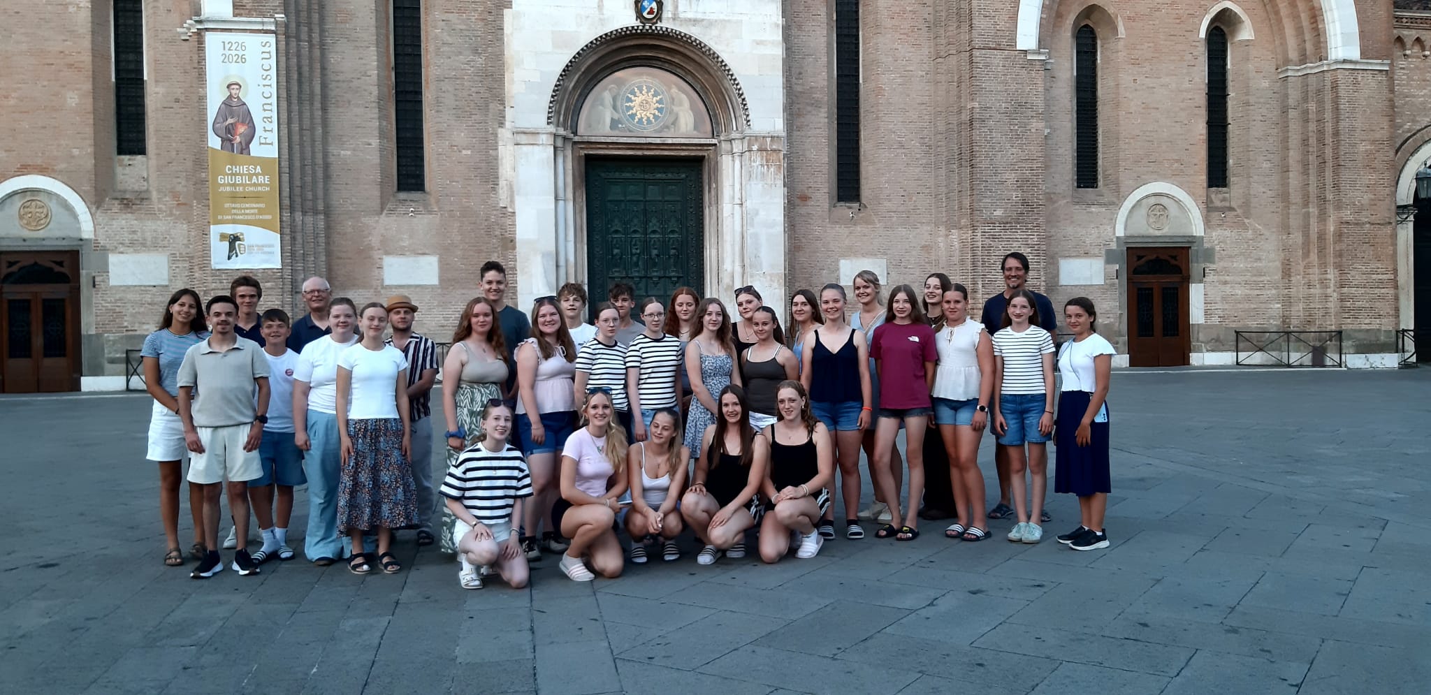Gruppenfoto mit fast ausschließlich Jugendlichen, die meisten in sommerlich kurzen Hosen und T-Shirts. Sie haben sich auf dem Platz vor einem runden Kirchenportal postiert, das offensichtlich zu einer größeren Anlage gehört: Rechts und links sind weitere Tore bzw. Türen zu sehen, dazwischen hohe, schmale Fenster, die z.T. oben abgeschnitten sind. An der Außenmauer der Kirche hängt eine hohes Transparent mit einer Heiligenfigur, auf dem steht: „1226 2026 Franciscus“ und „Chiesa giubilare“, dazu ein Tau.