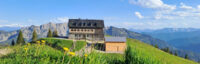 Auf einer grünen Anhöhe steht vor einer Bergkulisse unter blauem Himmel ein gemauertes Übernachtungshaus mit hölzernem Schuppen.
