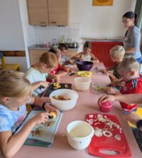 In einer Küche sitzen an einem langen Tisch Kinder und vor Schneidbrettern und bereiten Essen zu, es stehen verschiedene Gefäße auf dem Tisch.
