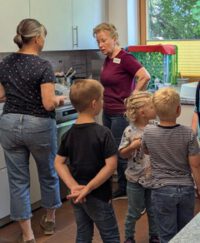 Zwei Frauen stehen in einer Küche an einer Arbeitszeile und sprechen miteinander. Davor stehen drei Kinder, die auf etwas außerhalb des Bildes blicken.