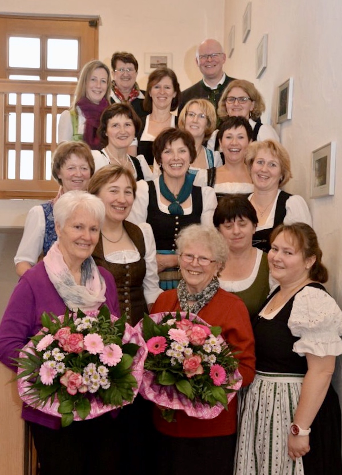 Gruppenfoto der Frauengemeinschaft Vogtareuth 2016: 15 Frauen in festlichen Dirndlkleidern haben sich auf einer Innentreppe aufgestellt und lachen in die Kamera. Ein einziger Mann steht ganz hinten/oben rechts (Pfarrer Guido Seidenberger). Zwei ältere Frauen ganz vorne halten große Blumensträuße in den Händen, die vorherrschende Farbe ist Rosa.