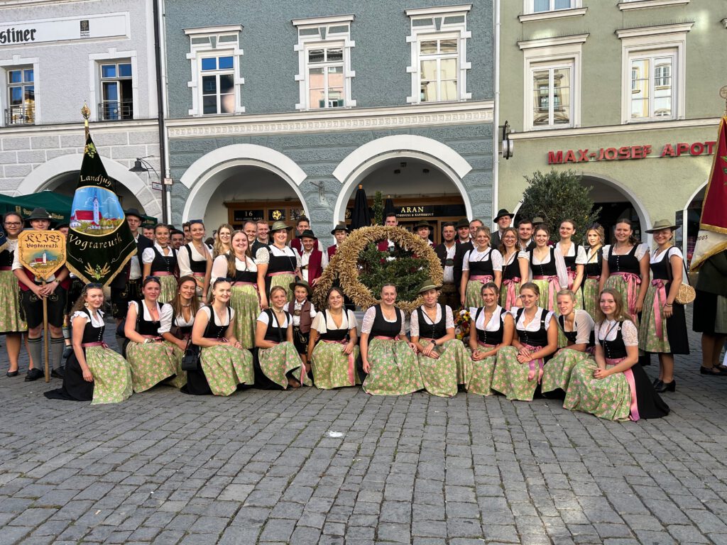 Auf einem Kopfsteinpflasterplatz haben sich vor den Arkaden von Altstadtfassaden junge Leute, vorwiegend Frauen, zum Gruppenbild postiert. Sie tragen einheitliche Tracht mit hellgrün gemusterten Schürzen, in der Mitte ist die Erntekrone. Am linken Rand trägt jemand eine grüne Fahne, auf der eine Kirche zu erkennen ist; auf der Fahne steht „Landjugend Vogtareuth“.Daneben trägt einer eine kleinere Tafel, ebenfalls mit Kirchenbild, auf der „KLJB Vogtareuth“ steht.