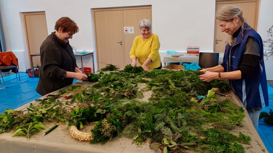 In einem Saal, der mit blauer Plane ausgelegt ist, stehen drei Frauen um einen großen Arbeitstisch, auf dem Nadelzweige und Kranz-Rohlinge sind, und fertigen Adventskränze.